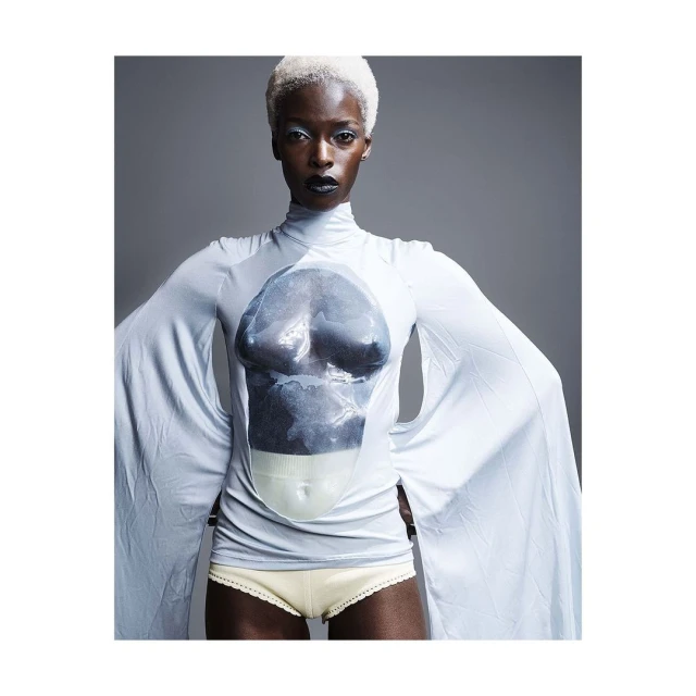 Striking studio portrait of a black female fashion model with white hair wearing a light blue long-sleeve top against gray backdrop.