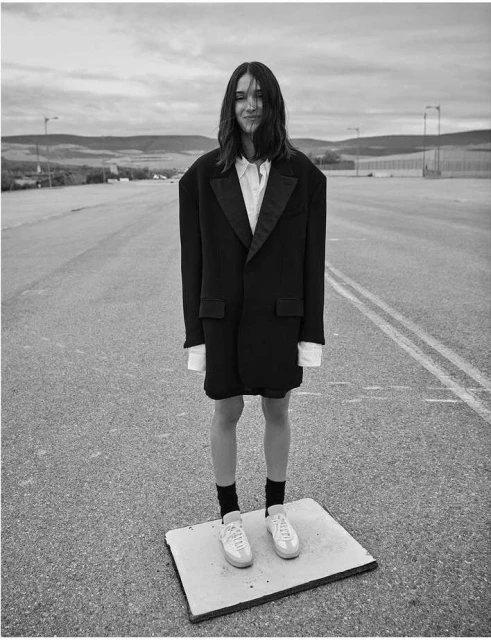 Black and white portrait of a young adult standing on a makeshift platform on a road, wearing an oversized black tuxedo jacket.