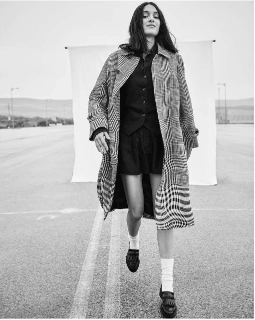 Woman in plaid coat and black dress walks towards the camera on a road with a white sheet backdrop in a black and white fashion shot.