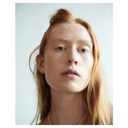Close-up portrait of a woman with red hair and light freckles wearing a neutral top against a plain wall.