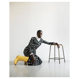 Black woman kneeling on a studio floor wearing a patterned black and white dress and yellow rubber boots