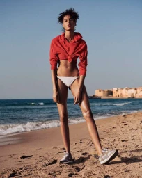 Model standing on a sandy beach wearing a red tied hoodie, white bikini bottoms, and sneakers, photographed by Jan Welters