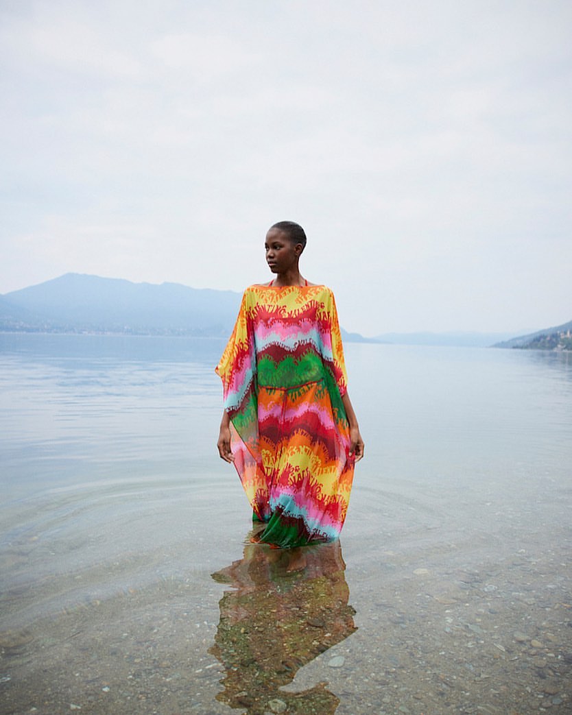 Fashion shot of an African woman standing in water wearing a colorful dress with lake and mountain background on an overcast day.