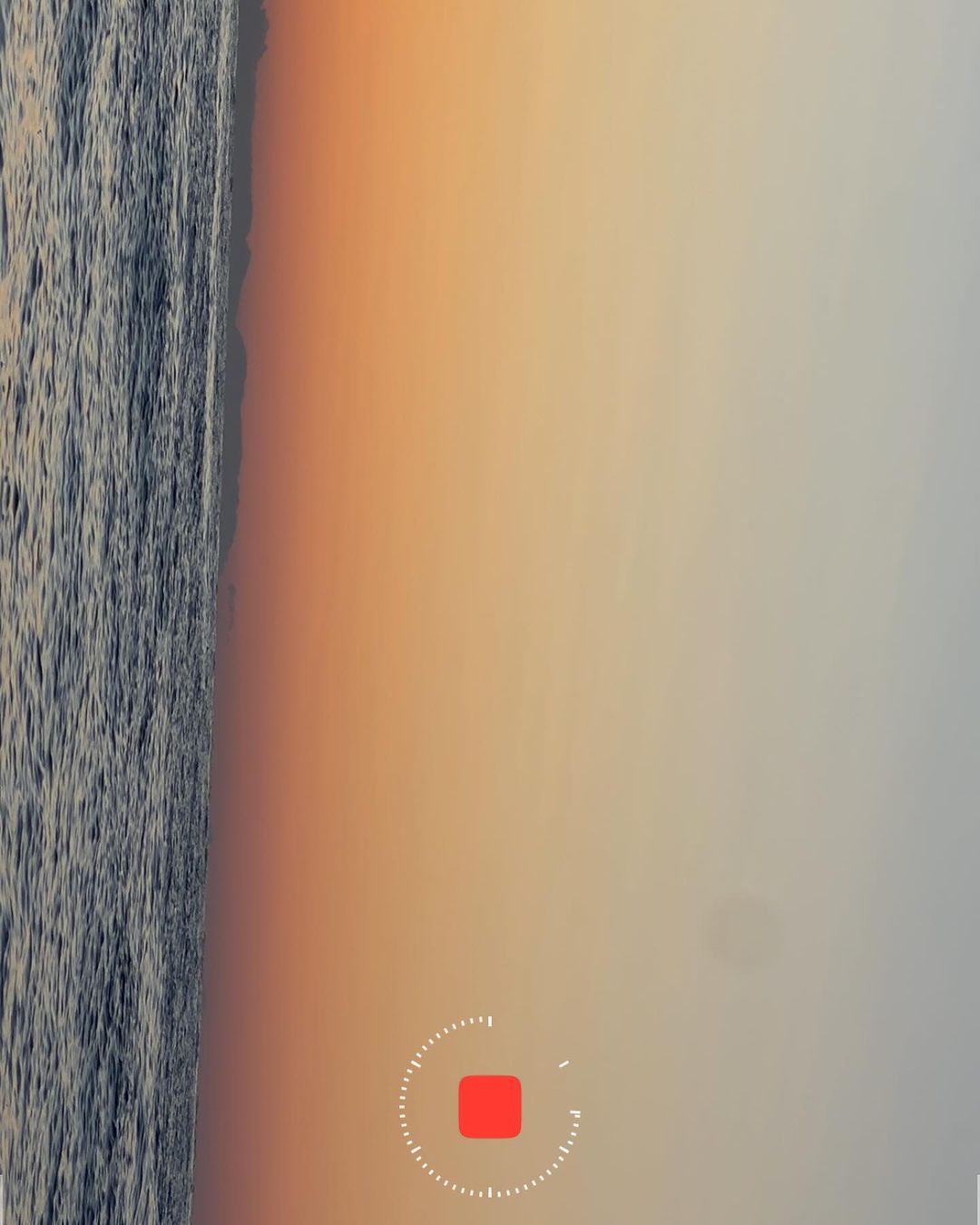 Overhead shot of the ocean meeting the sandy shore at dusk, the sky a blend of orange and light grey, captured with a stationary recording indicator.