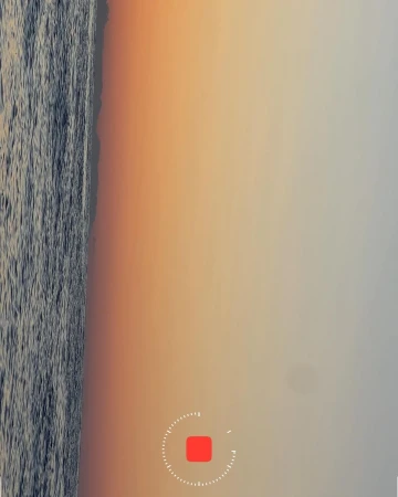 Overhead shot of the ocean meeting the sandy shore at dusk, the sky a blend of orange and light grey, captured with a stationary recording indicator.