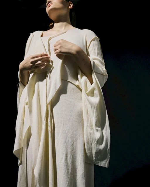 Fashionable woman in flowing beige dress, standing in a dimly lit setting with a mix of direct and diffused light creating soft shadows.