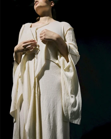 Fashionable woman in flowing beige dress, standing in a dimly lit setting with a mix of direct and diffused light creating soft shadows.