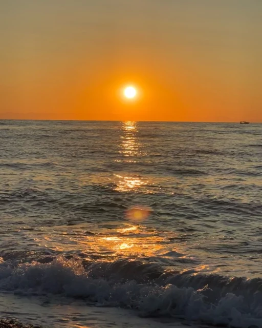 Golden sunset over the ocean. Waves gently roll onto the shore, reflecting sunlight. A small boat sits on the horizon under the warm sky.