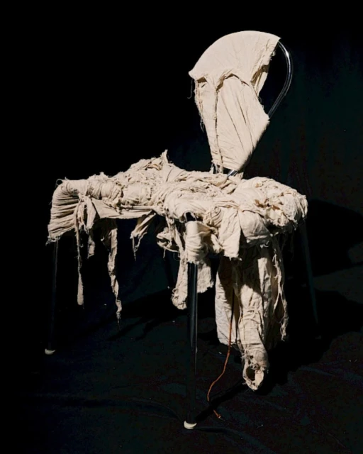 Artistic photo of a chair draped in tattered beige cloth, creating a draped effect, set against a dark background in a studio setting.