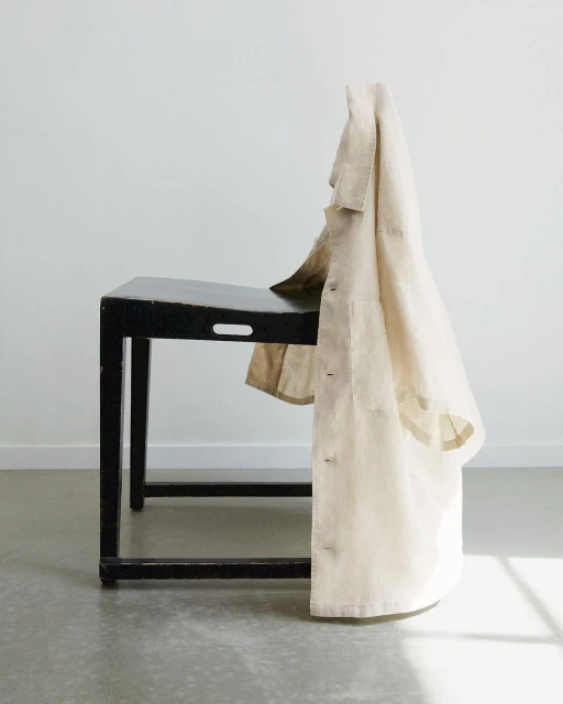 Beige linen lab coat drapes over a dark wood side table against a plain white background, lit by natural light in a minimal aesthetic.