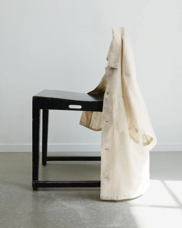Beige linen lab coat drapes over a dark wood side table against a plain white background, lit by natural light in a minimal aesthetic.