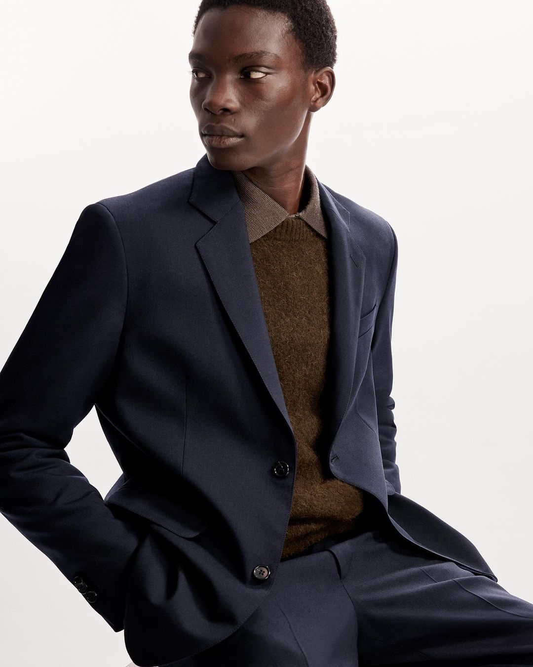 An adult male model poses indoors wearing a tailored navy blue suit and brown collared sweater in a studio setting.