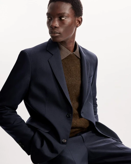 An adult male model poses indoors wearing a tailored navy blue suit and brown collared sweater in a studio setting.