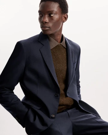 An adult male model poses indoors wearing a tailored navy blue suit and brown collared sweater in a studio setting.