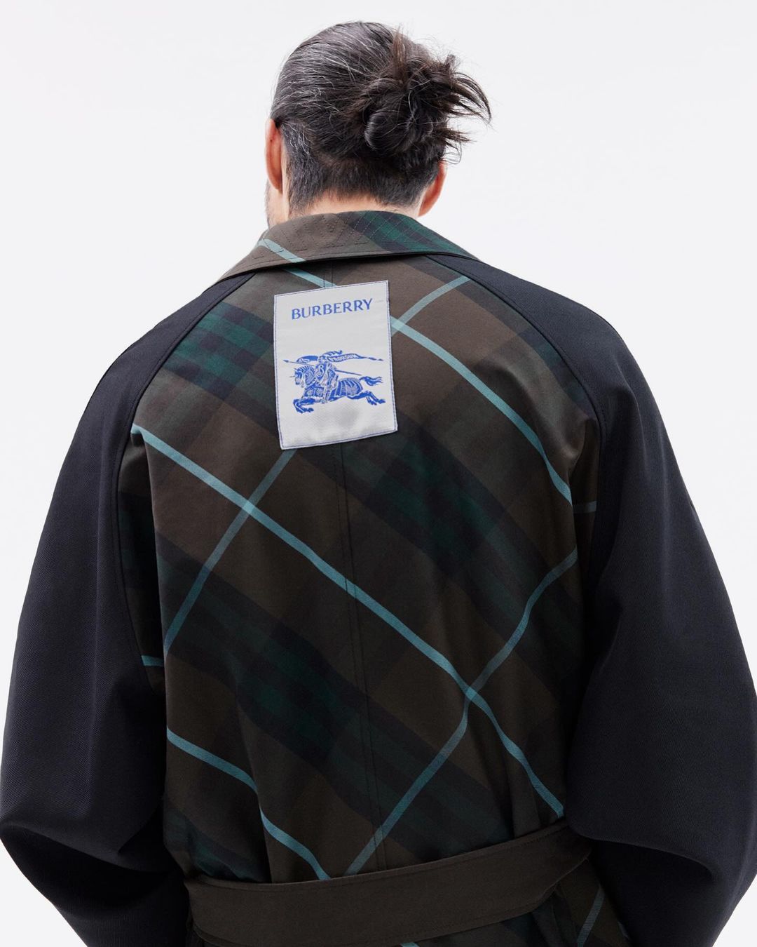 An adult male from the rear wearing a black and green plaid coat with a Burberry logo patch on the upper back against a white backdrop.