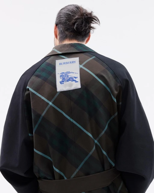 An adult male from the rear wearing a black and green plaid coat with a Burberry logo patch on the upper back against a white backdrop.