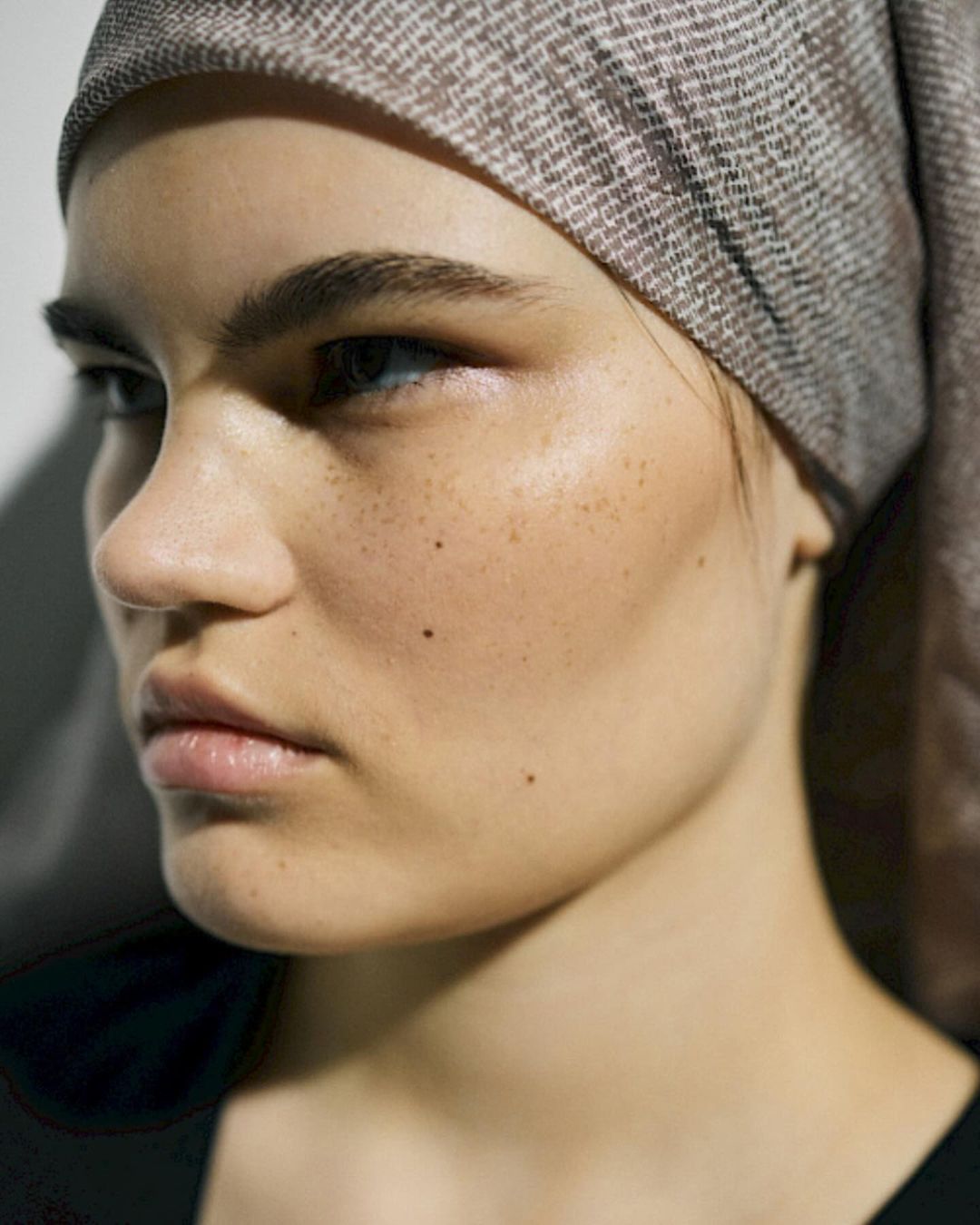 Close-up portrait of a young woman with freckles, wearing a patterned headscarf, and looking off to the side in a brightly lit studio.