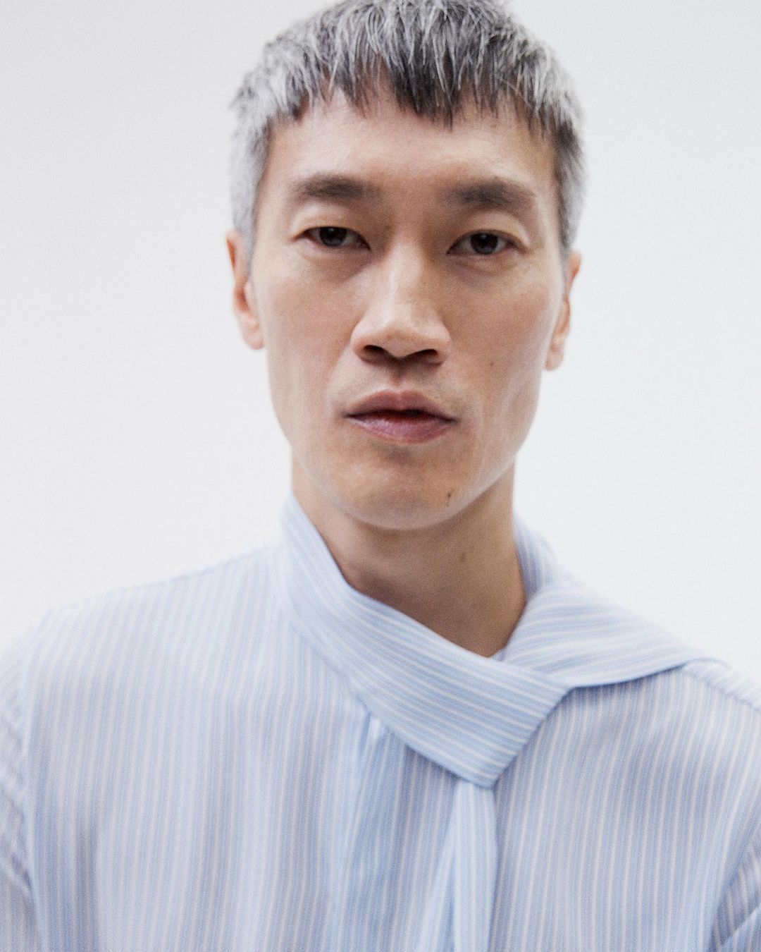 Close-up studio portrait of an Asian male model with silver hair wearing a striped shirt with a tied collar against a white backdrop.