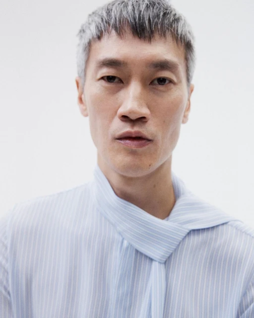 Close-up studio portrait of an Asian male model with silver hair wearing a striped shirt with a tied collar against a white backdrop.