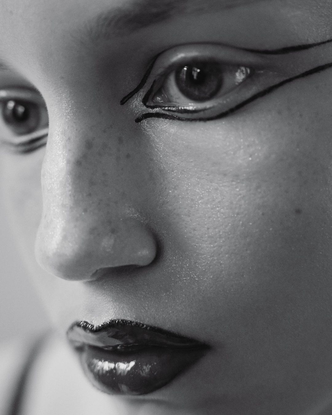 Close-up black and white portrait of a person's face featuring glossy dark lipstick and distinct black graphic eyeliner.
