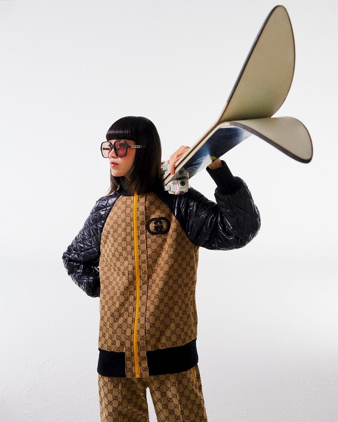 Woman in Gucci tracksuit poses with snowboard on white background in studio shot, accessorized with sunglasses and black quilted jacket.