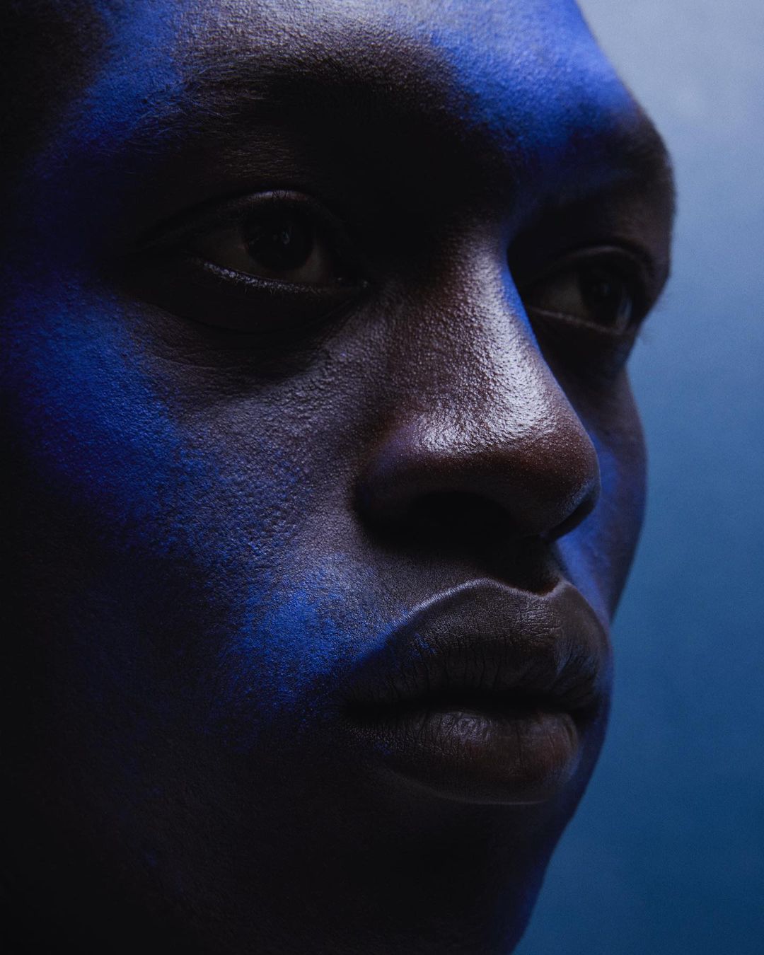 Close-up studio portrait of a Black man with dark skin illuminated with blue makeup, creating a high-contrast, artistic aesthetic.