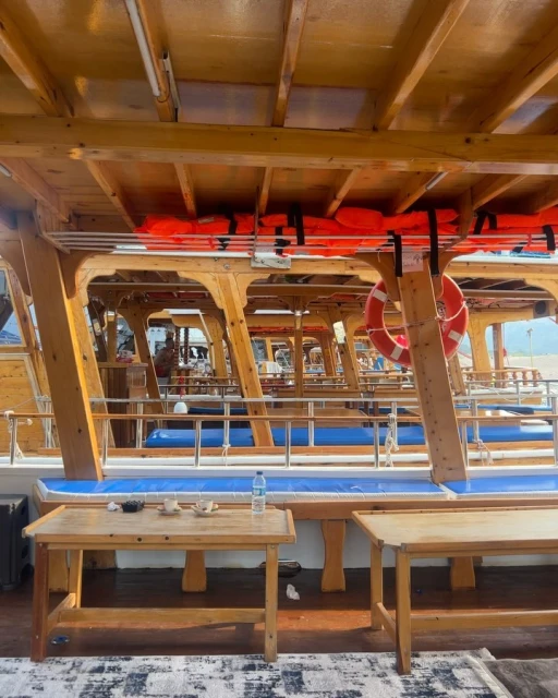Interior view of a boat featuring wooden tables and benches with safety equipment like life jackets, under a wooden ceiling.