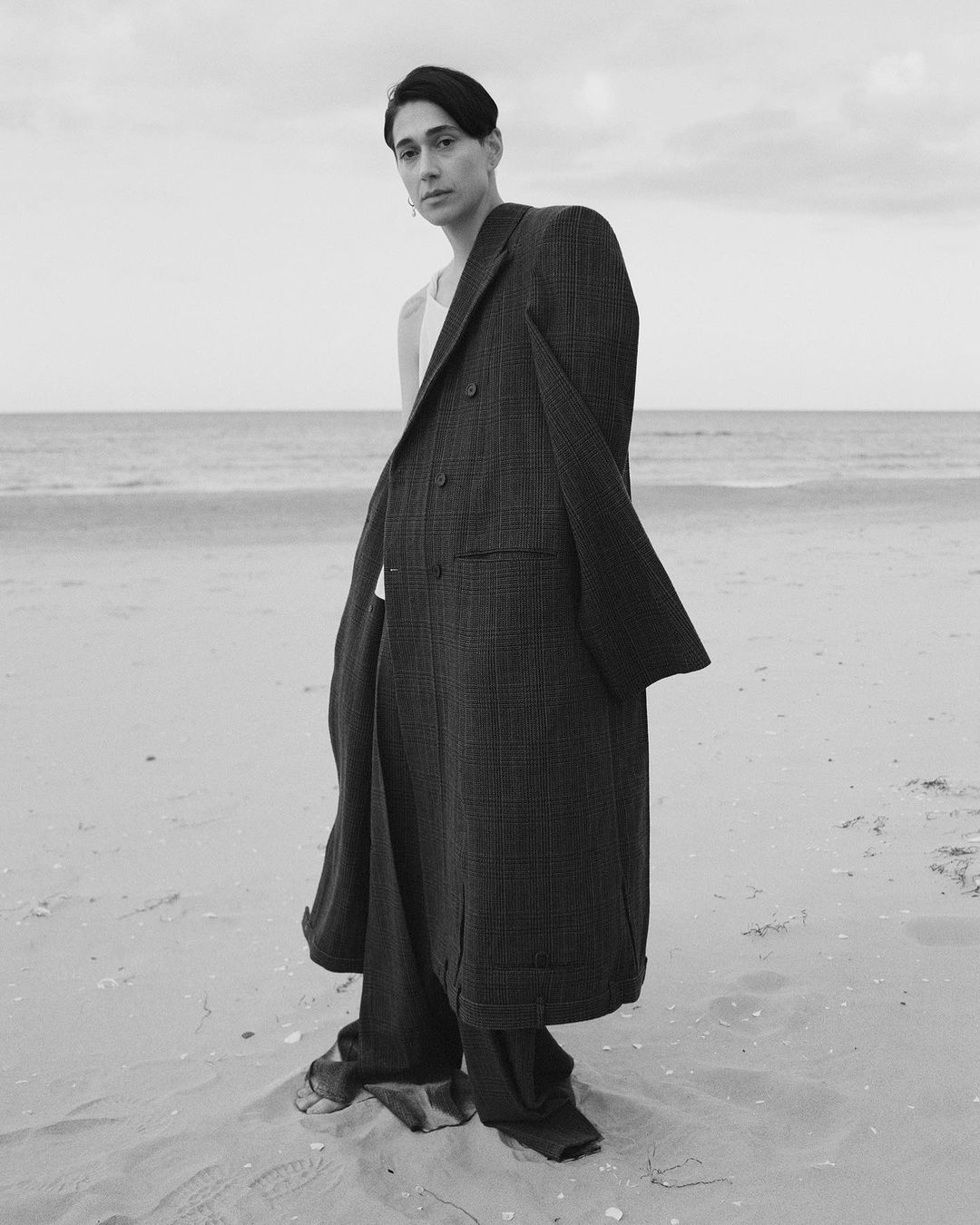 Monochrome image of an adult in an oversized coat standing on a beach with a sea background under a cloudy sky.