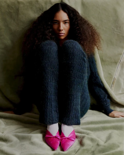 Portrait of a brunette woman with curly hair wearing a dark sweater and pink shoes sitting on a textured green surface.