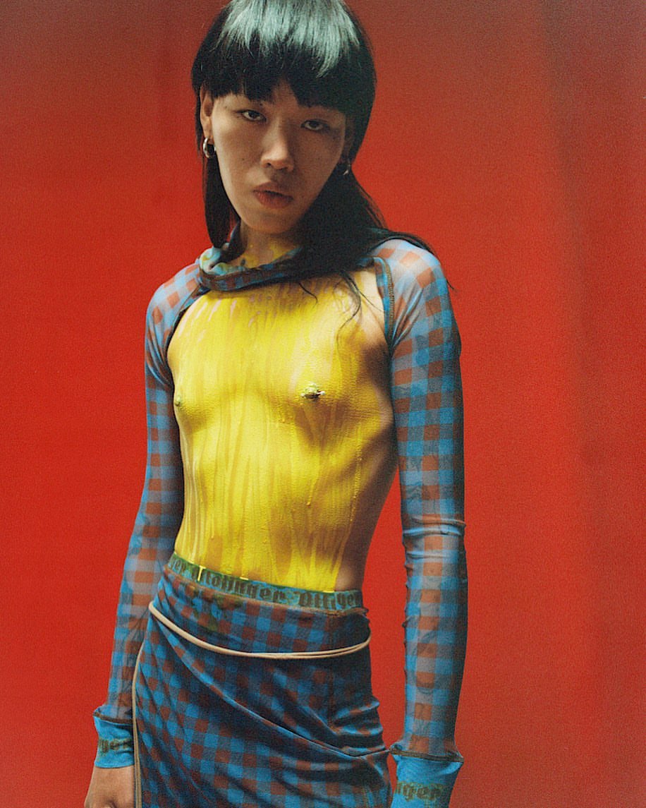 Portrait of a person with dark hair, wearing a blue and brown patterned top and skirt, against a red studio backdrop.