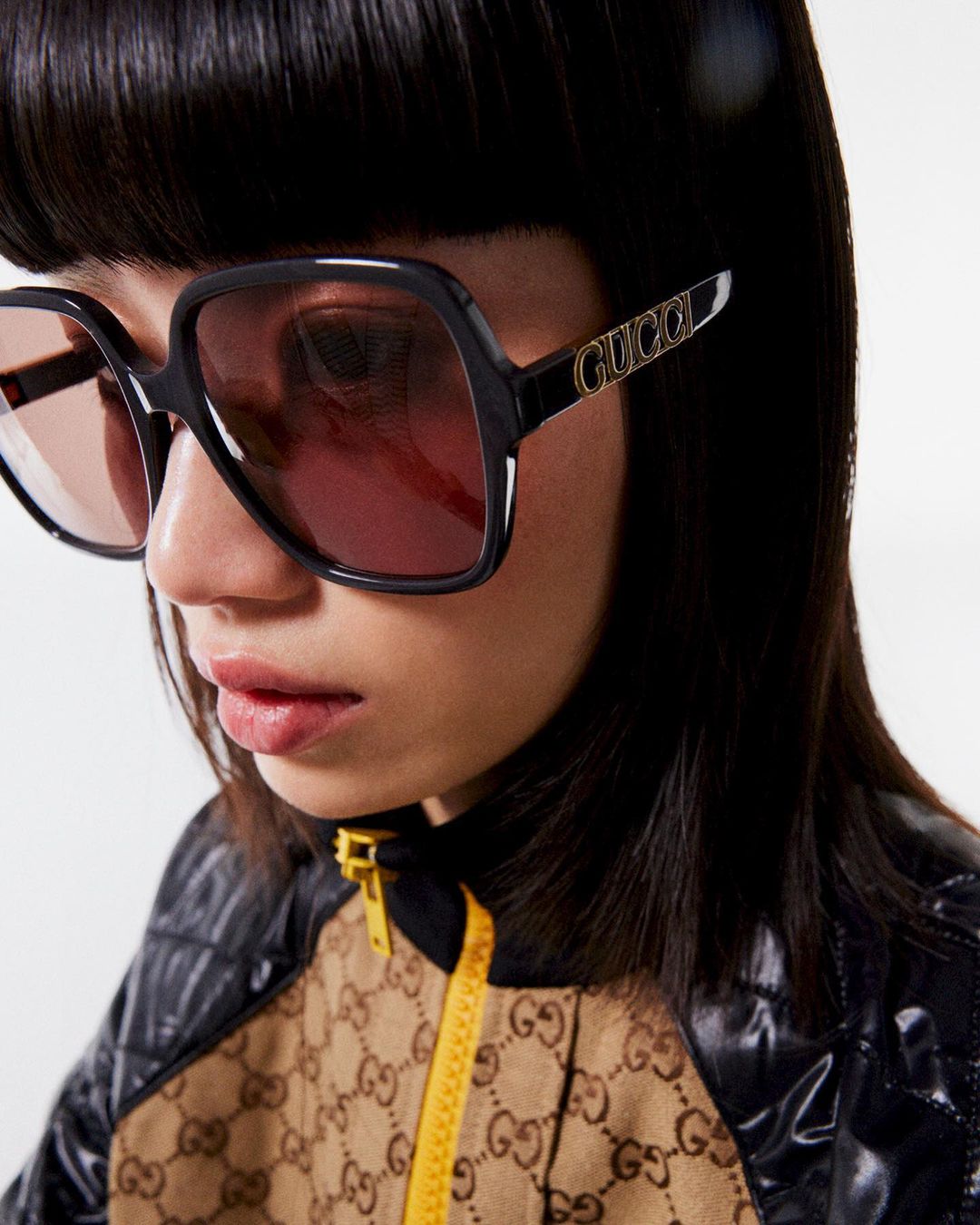 A woman with dark hair and black-framed Gucci sunglasses looks directly at the camera, modeling fashion apparel in an indoor setting.