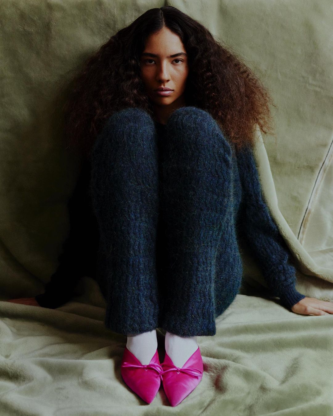 A woman with dark, curly hair is seated indoors with a neutral background, wearing dark blue pants, a matching top, and pink shoes.