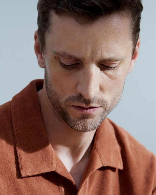 Close-up portrait of an adult man with fair skin, light stubble, and an orange shirt, set against a plain, light background.