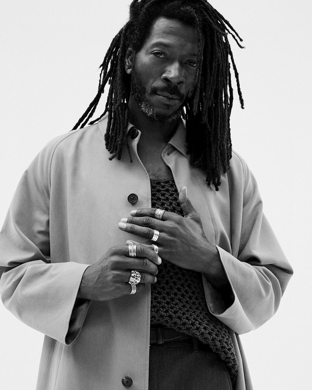 Black male model with dreadlocks wearing a tan jacket and rings against a white background in a studio portrait with eye-level camera angle.