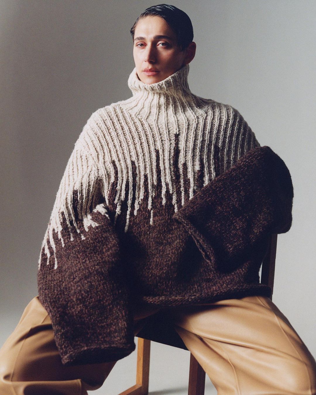 Woman wearing a large brown and beige turtleneck sweater posing indoors. She is sitting on a wooden chair, and wearing tan-colored pants.