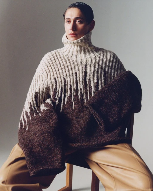 Woman wearing a large brown and beige turtleneck sweater posing indoors. She is sitting on a wooden chair, and wearing tan-colored pants.