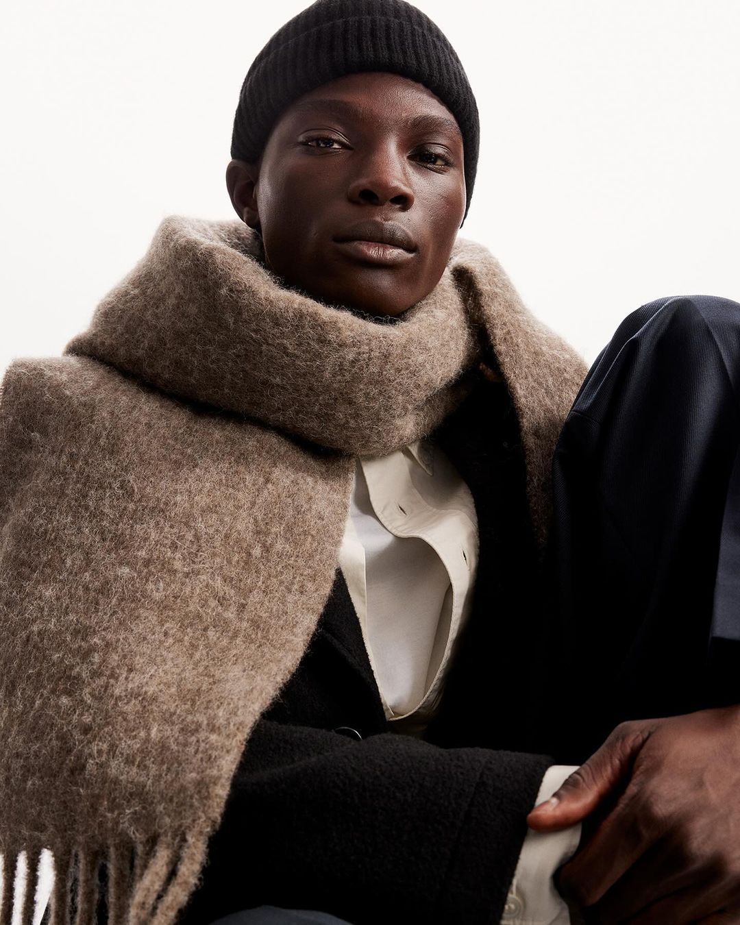 Portrait of a Black male wearing a black beanie and a large wool scarf, staring directly at the camera against a neutral background.