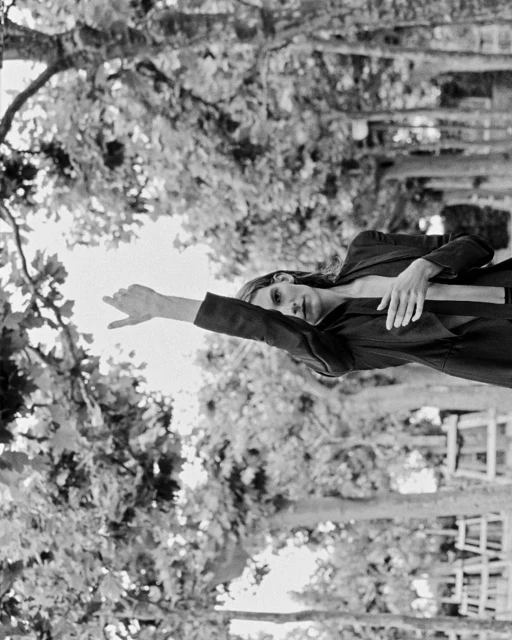 Black and white photograph of a woman in a blazer outside, reaching one arm toward the sky, among the trees and sunlight.