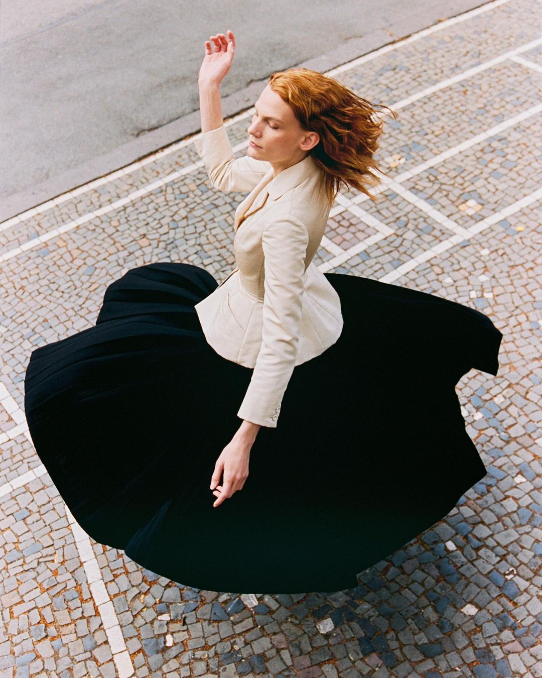 Woman in beige blazer and black dress dances on mosaic sidewalk, elevated view, capturing movement and fashion.