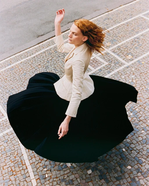 Woman in beige blazer and black dress dances on mosaic sidewalk, elevated view, capturing movement and fashion.
