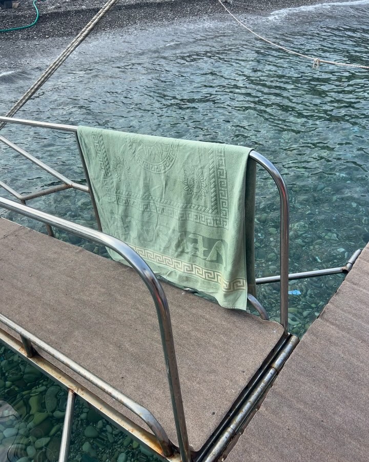 View of a light green towel hanging on a metal rail near a rocky beach and clear turquoise water on a sunny day.