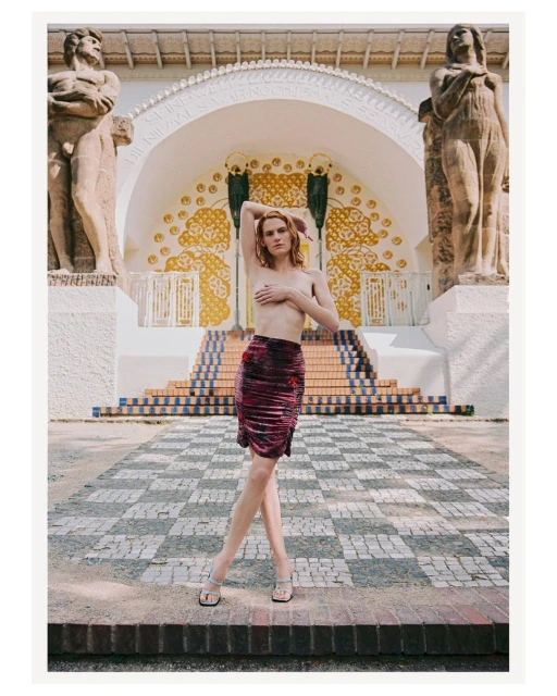 Nude woman in a skirt poses in front of art nouveau architecture and statues on a checkered stone plaza in Vienna, Austria.