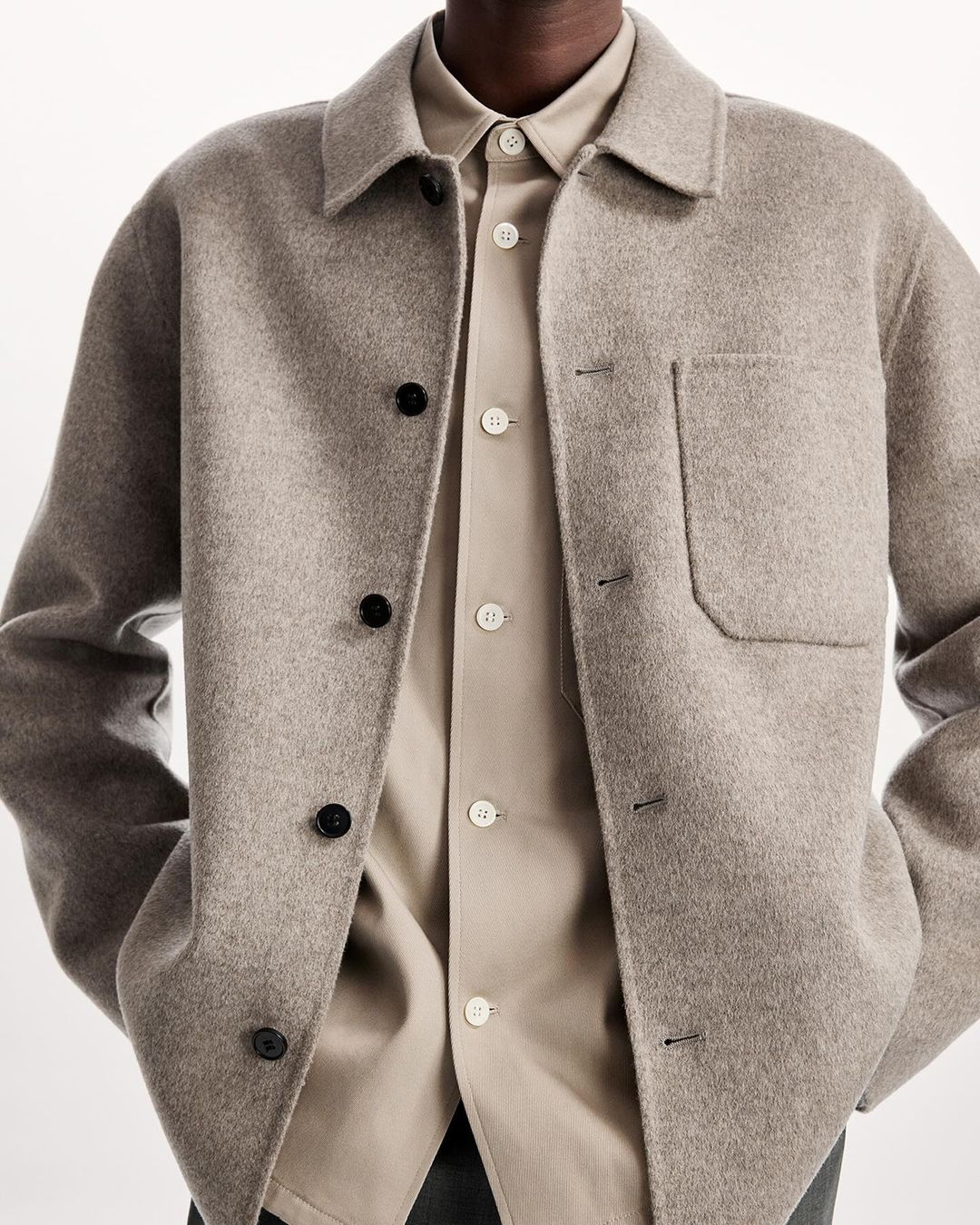 Close-up of a grey wool overshirt, layered over a beige button-down shirt, featuring black buttons on the overshirt and light buttons on the shirt.