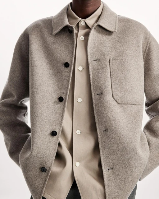 Close-up of a grey wool overshirt, layered over a beige button-down shirt, featuring black buttons on the overshirt and light buttons on the shirt.