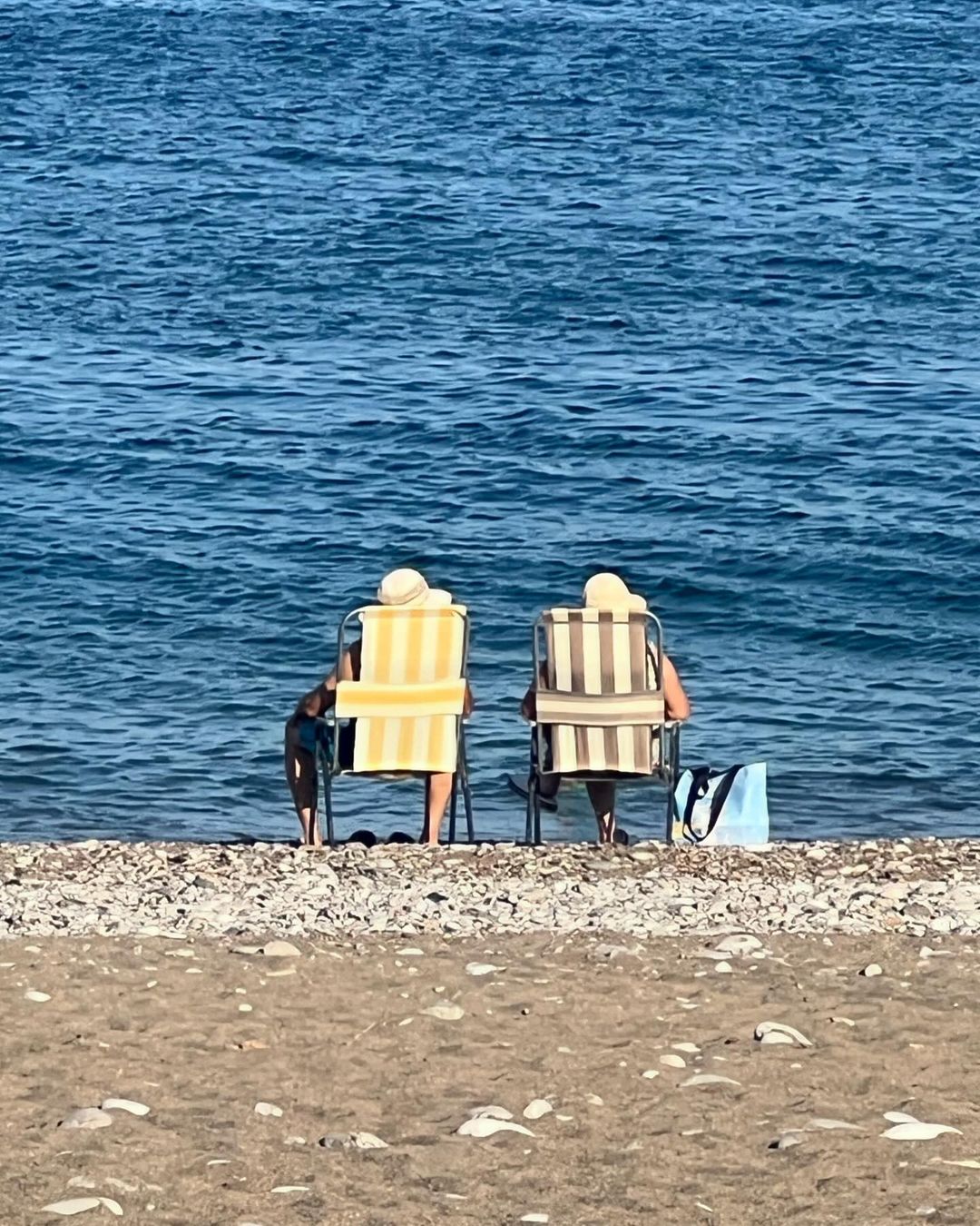 Two people sitting on beach chairs by the ocean with a dog and a tote bag on a sunny day, enjoying the coastal scenery.