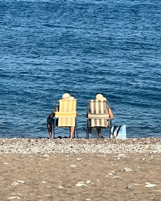 Two people sitting on beach chairs by the ocean with a dog and a tote bag on a sunny day, enjoying the coastal scenery.