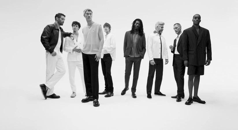 Black and white studio shot of group of eight men in smart casual outfits including jackets, suits, shirts, and dress shoes.