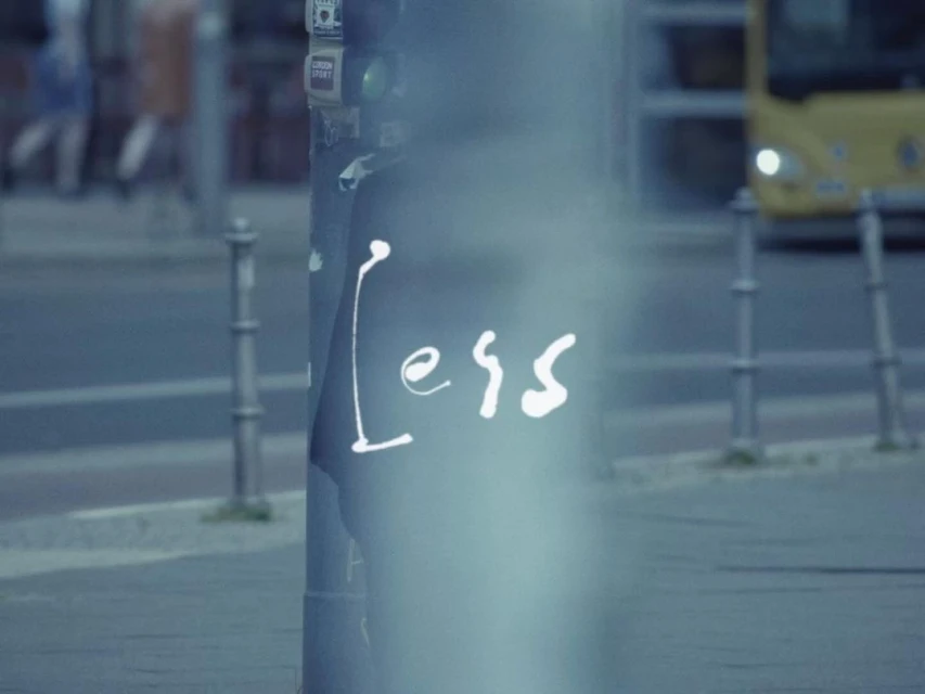 Urban scene with a weathered signpost displaying the handwritten word 'Less' in white against a blurred cityscape backdrop and a bus.