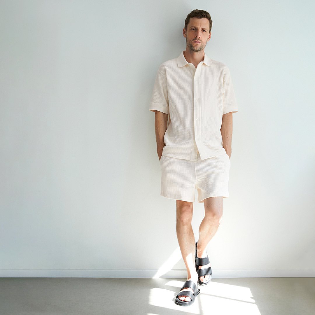 Man in matching cream-colored shirt and shorts stands against a white wall in a casual pose wearing black sandals. Minimalist fashion.