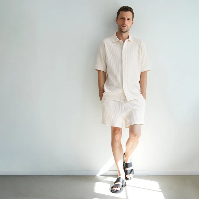 Man in matching cream-colored shirt and shorts stands against a white wall in a casual pose wearing black sandals. Minimalist fashion.